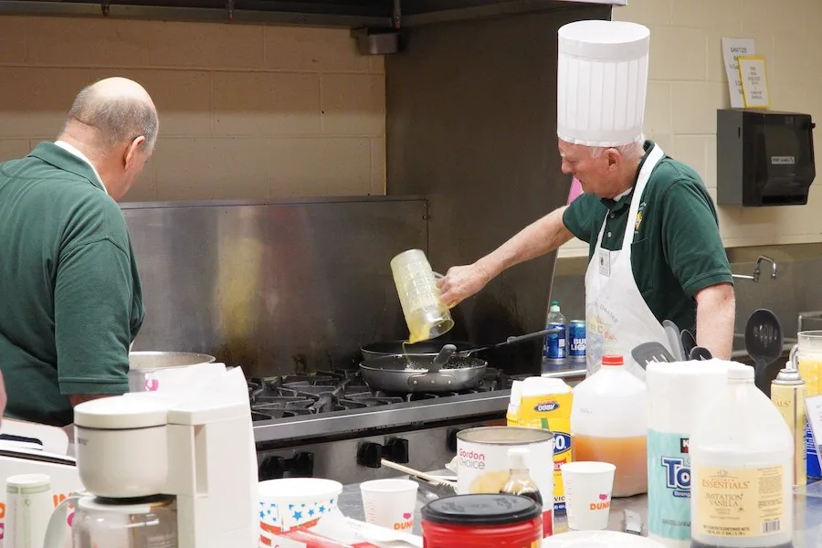 Two council members at the parish kitchen stove, one pouring batter into a pan